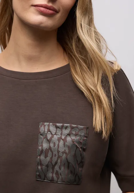 Frau in schlichtem, braunem T-Shirt mit gemusterter, lederähnlicher Brusttasche.